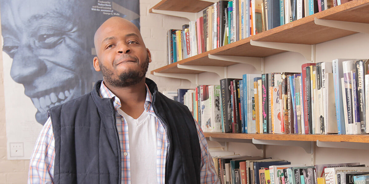 Photo of Kiese Laymon standing in front of a photo of James Baldwin and next to a bookshelf
